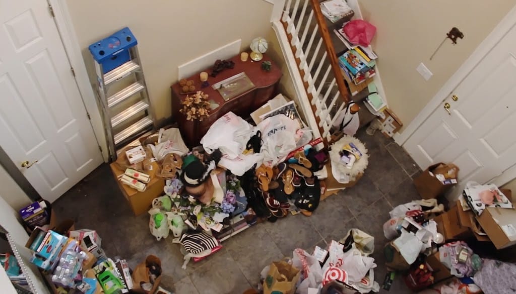 An overhead view of a heavily cluttered home entryway and staircase filled with piles of bags, boxes, shoes, and household items.