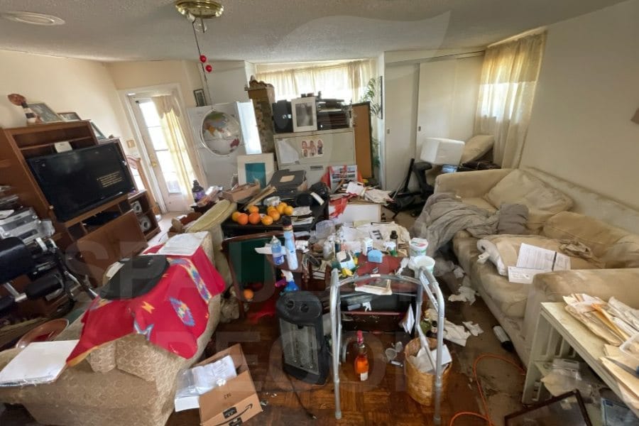 A cluttered living room with scattered papers, boxes, and household items on the floor and furniture. Shelves, a TV, and covered couches are visible amid the mess. 