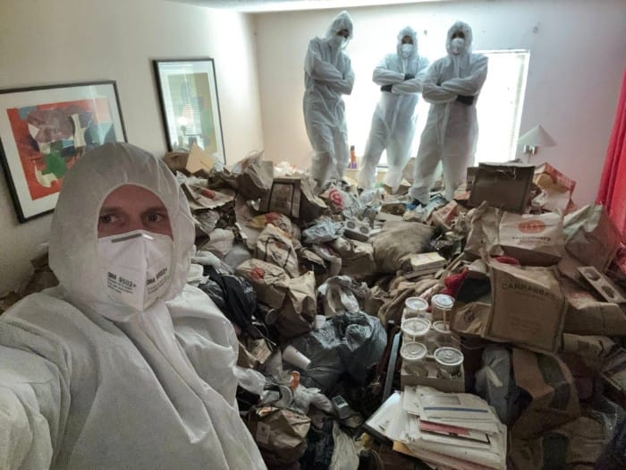 A cleanup team in full biohazard suits and masks inside a room filled with extreme hoarding, primarily consisting of takeout bags